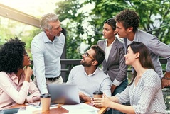 Grupo de seis pessoas discutindo e colaborando ao ar livre, envolvendo um gerente com equipe diversificada, em um ambiente profissional ao ar livre, com árvores verdes ao fundo e um laptop na mesa.