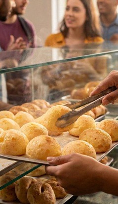 Atendente usando avental serve pães de queijo fresquinhos com uma pinça, organizando-os em uma bandeja atrás do balcão de vidro. Do outro lado da vitrine, vários clientes aguardam em fila no ambiente movimentado de uma padaria ou lanchonete.