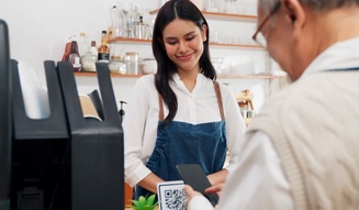 Jovem mulher sorridente fazendo pagamento com smartphone em uma cafeteria, usando uniforme e atendente ao lado, ambiente acolhedor e bem decorado