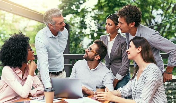 Grupo de seis pessoas discutindo e colaborando ao ar livre, envolvendo um gerente com equipe diversificada, em um ambiente profissional ao ar livre, com árvores verdes ao fundo e um laptop na mesa.